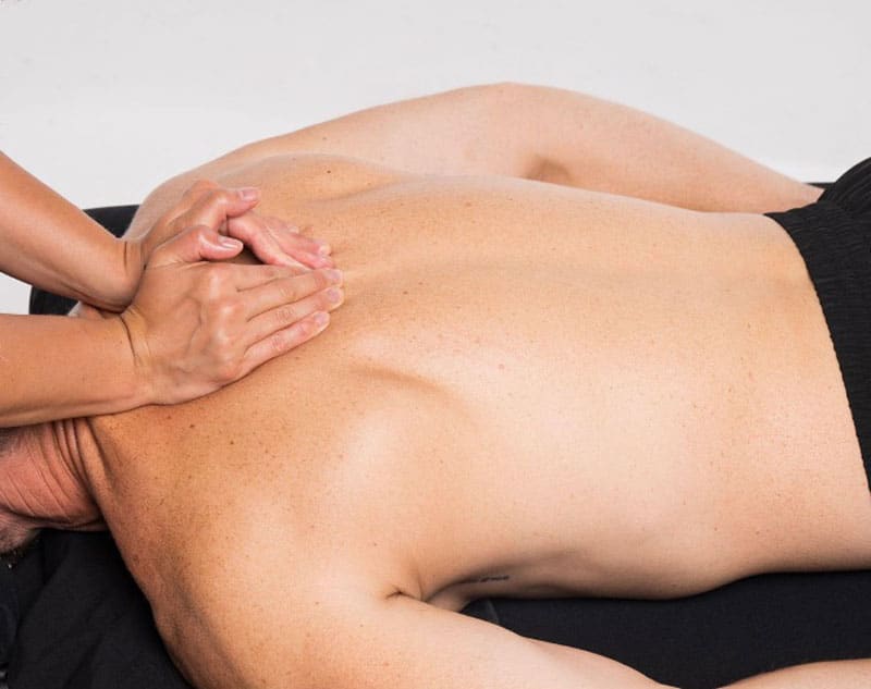 A person is receiving a back massage while lying face down. Another person's hands are seen applying pressure to the upper back.