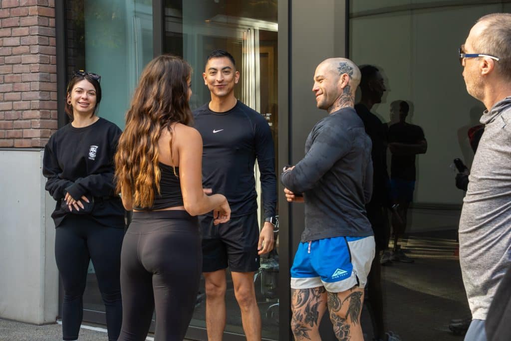 A group of five people stand chatting outside a building. One person has tattoos, and another wears a black long-sleeve top. They appear relaxed and engaged in conversation.