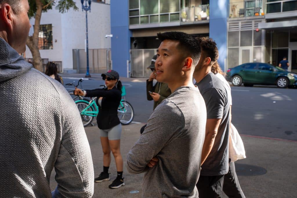 A group of people stand on a city sidewalk. A man in a gray shirt faces others. A woman with a cap and sunglasses points at something. A bicycle and car are in the background.
